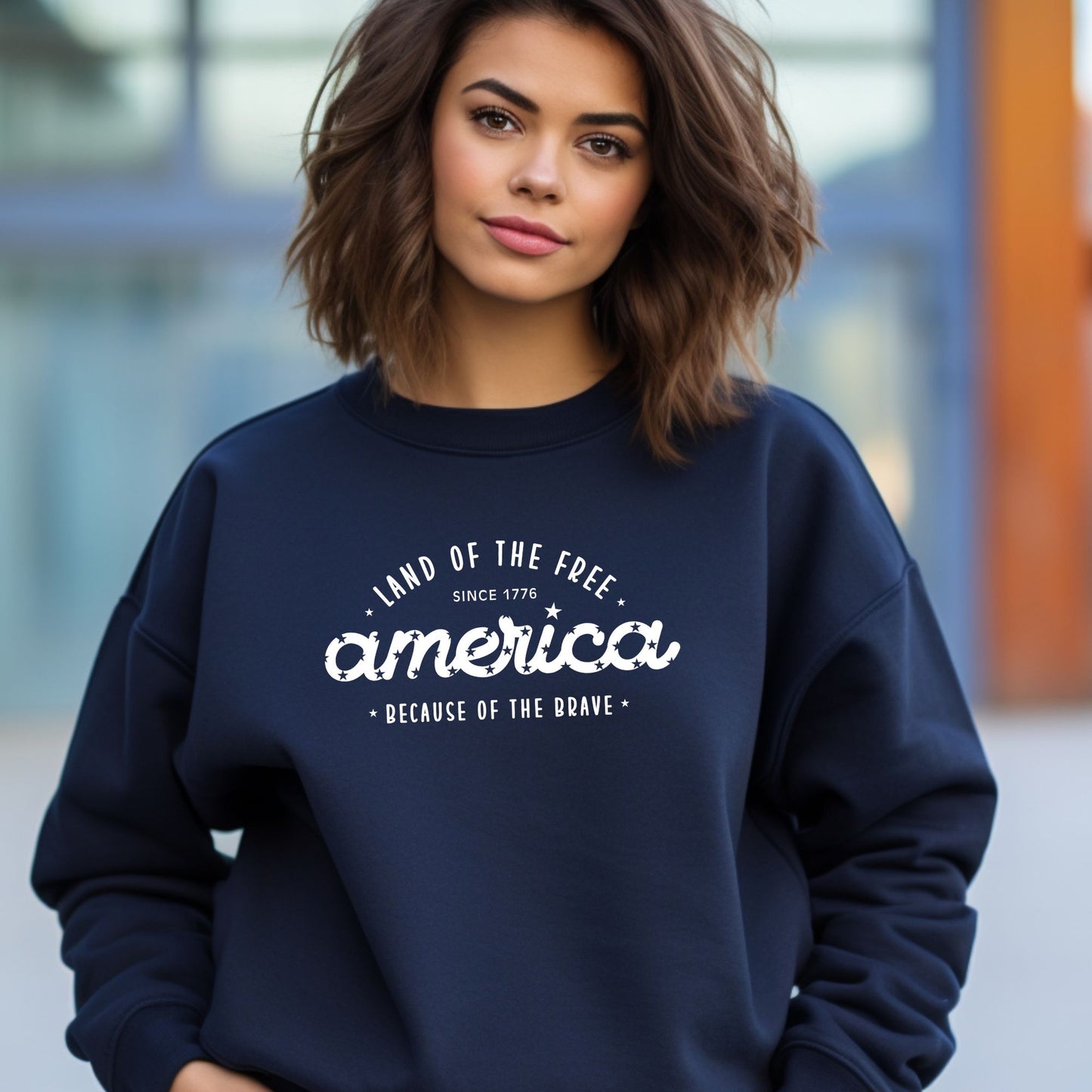 Woman wearing a navy patriotic crewneck that says "Land of the Free, America, Because of the Brave.