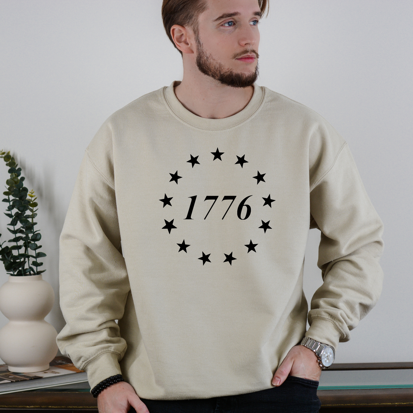 1776 circle stars graphic on sand-colored crewneck sweatshirt, modeled indoors with minimalist decor.