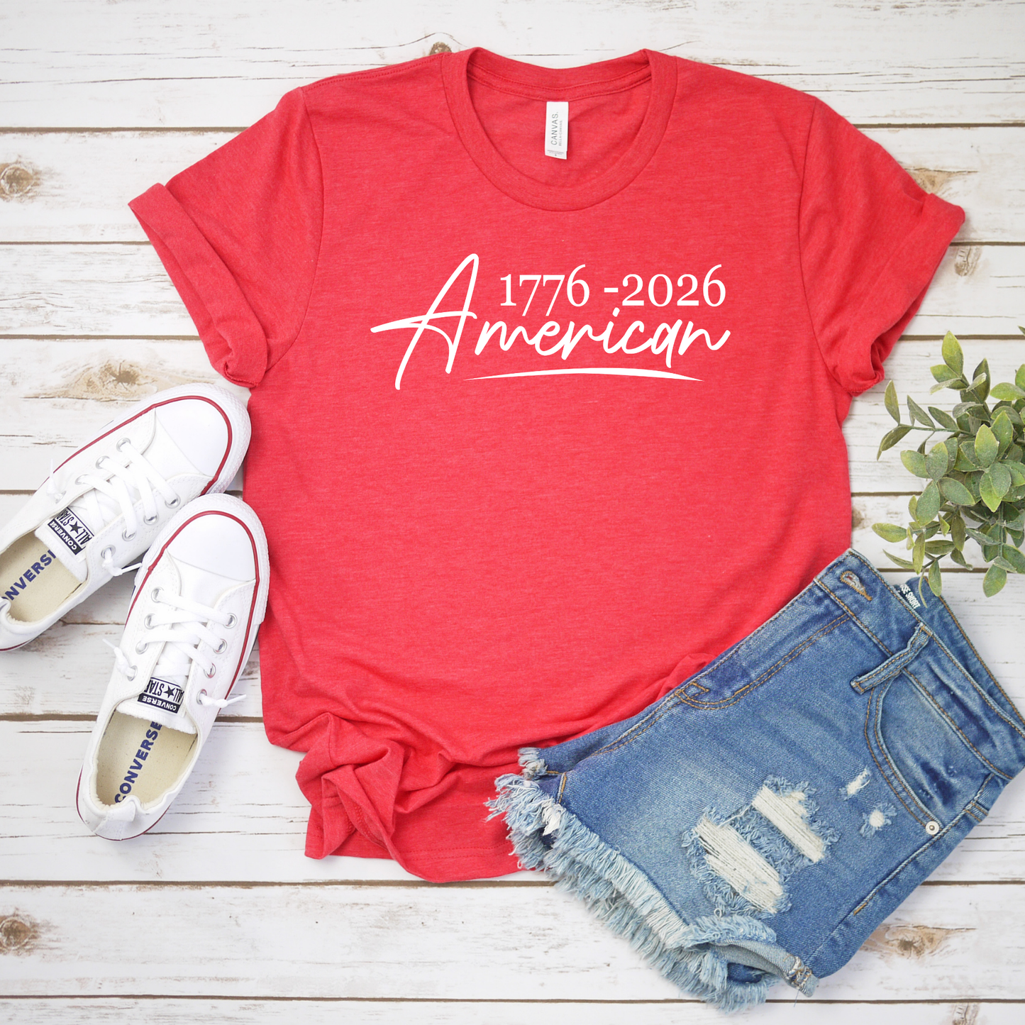 Red t-shirt with '1776-2026 American' text, white sneakers, and denim shorts on a wooden surface.