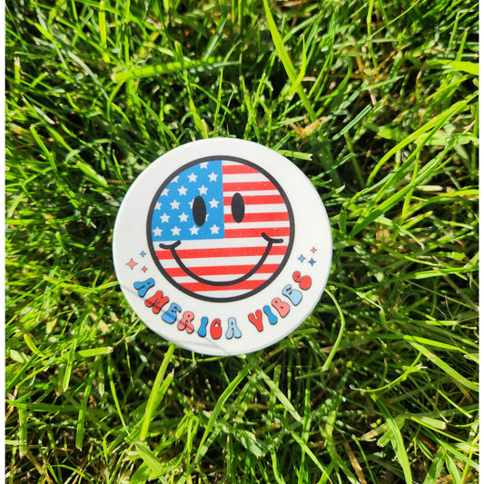 Patriotic America Vibes sticker with red, white, and blue smiley face design laying on vibrant green grass.