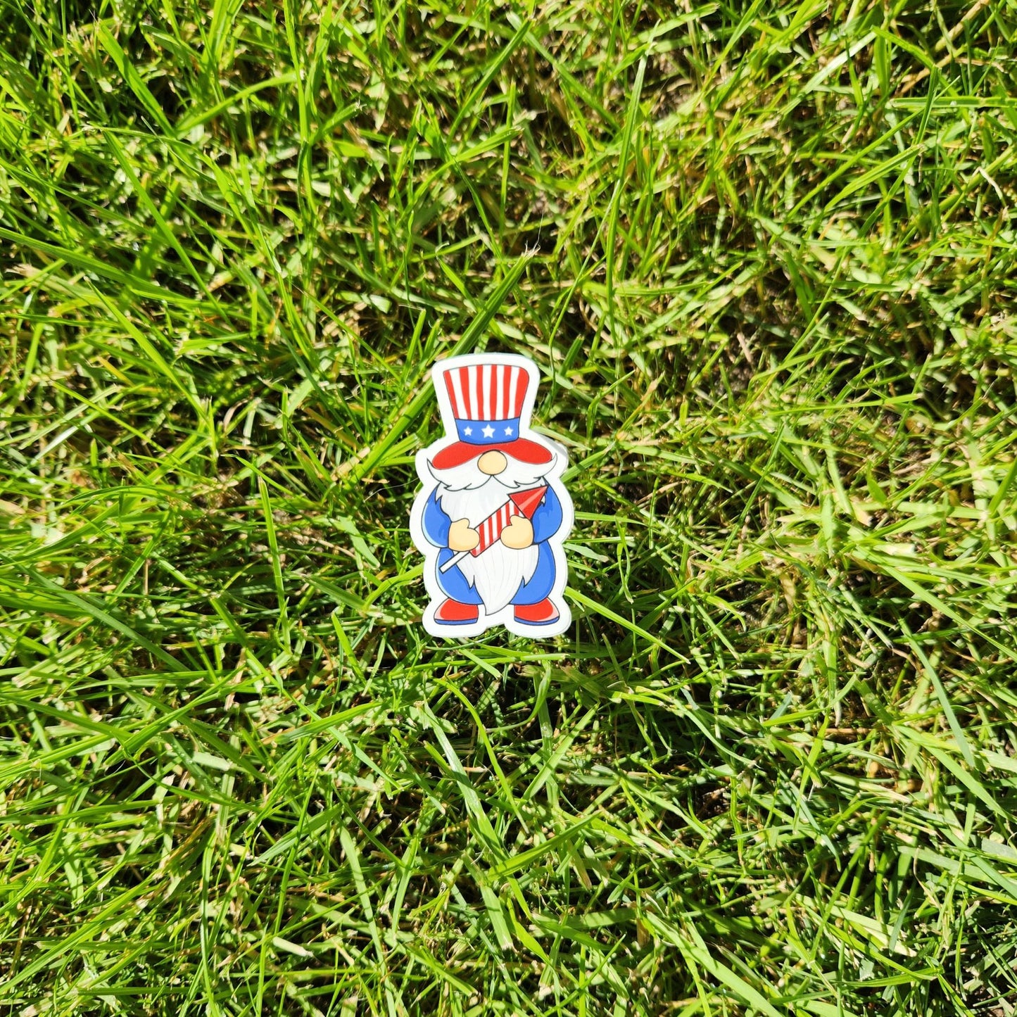Patriotic gnome sticker in red, white, and blue with firework rocket, placed on grass.