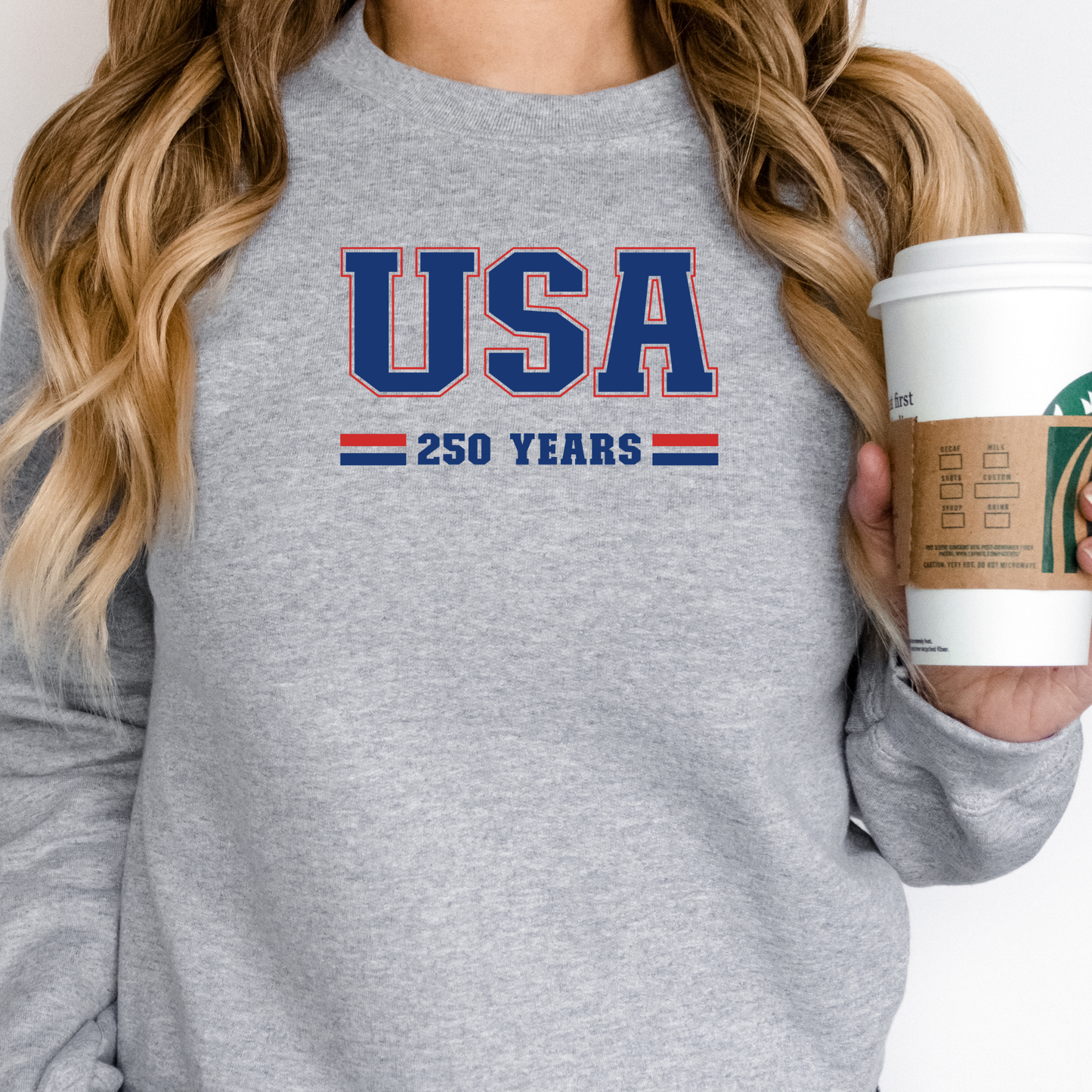Person wearing a gray sweatshirt with 'USA 250 Years' text, holding a Starbucks coffee cup.