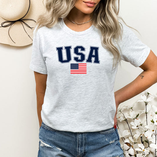 Woman modeling a light ash gray USA t-shirt with navy lettering and American flag, styled with denim shorts for a summer-ready patriotic outfit.