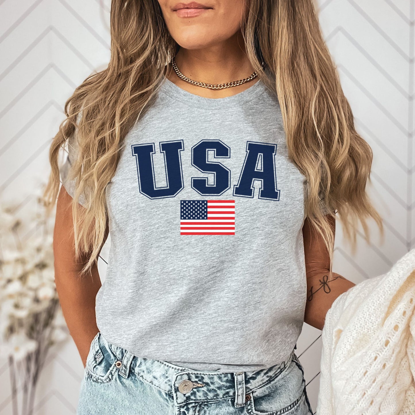 Woman in an athletic gray USA t-shirt with bold navy text and American flag, styled casually with high-waisted denim shorts.