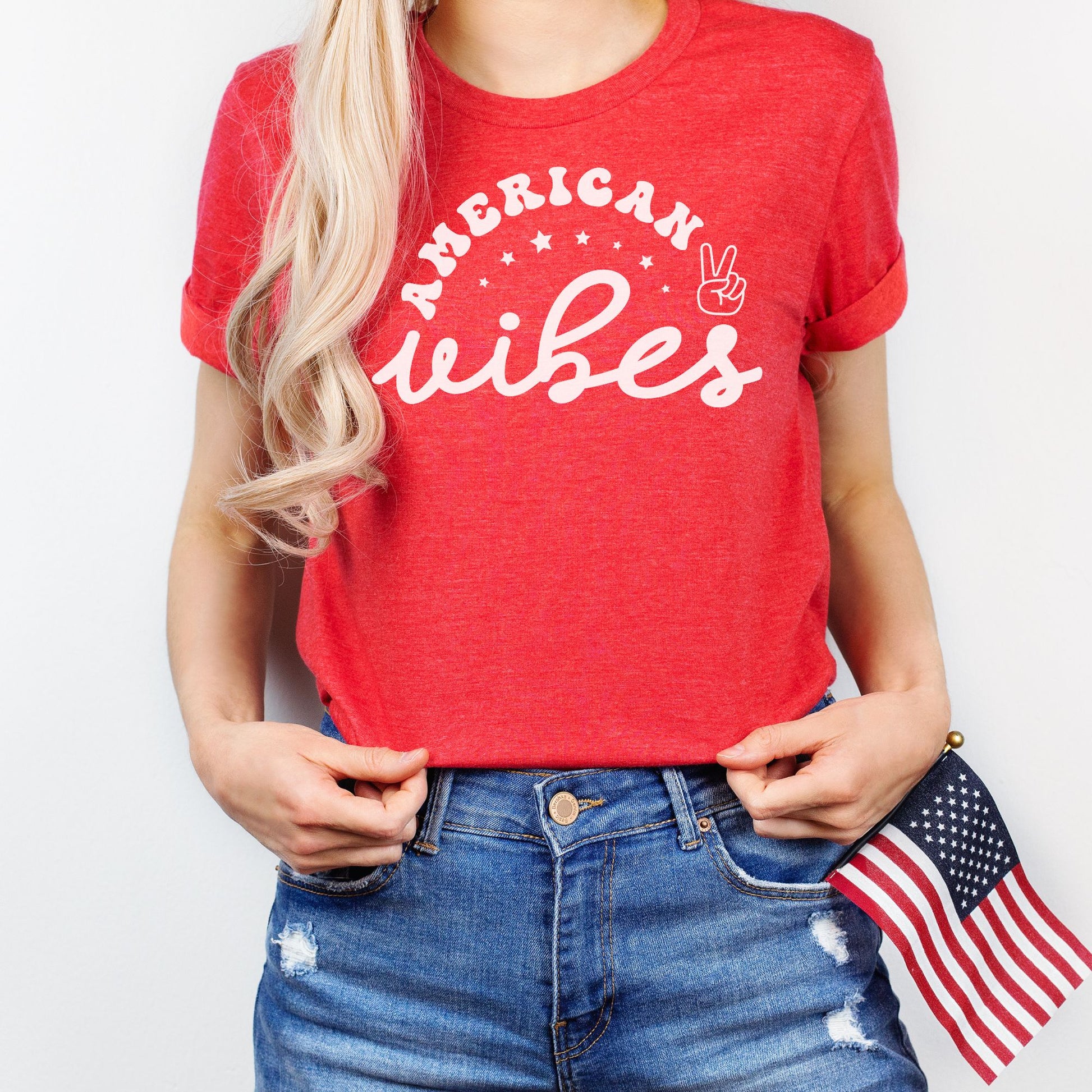 Patriotic graphic tee in heather red featuring "American Vibes" with stars and peace sign; woman holding a small American flag.
