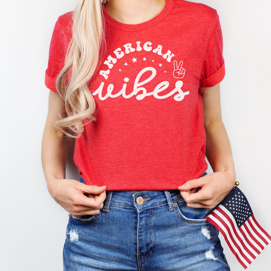 Patriotic graphic tee in heather red featuring "American Vibes" with stars and peace sign; woman holding a small American flag.