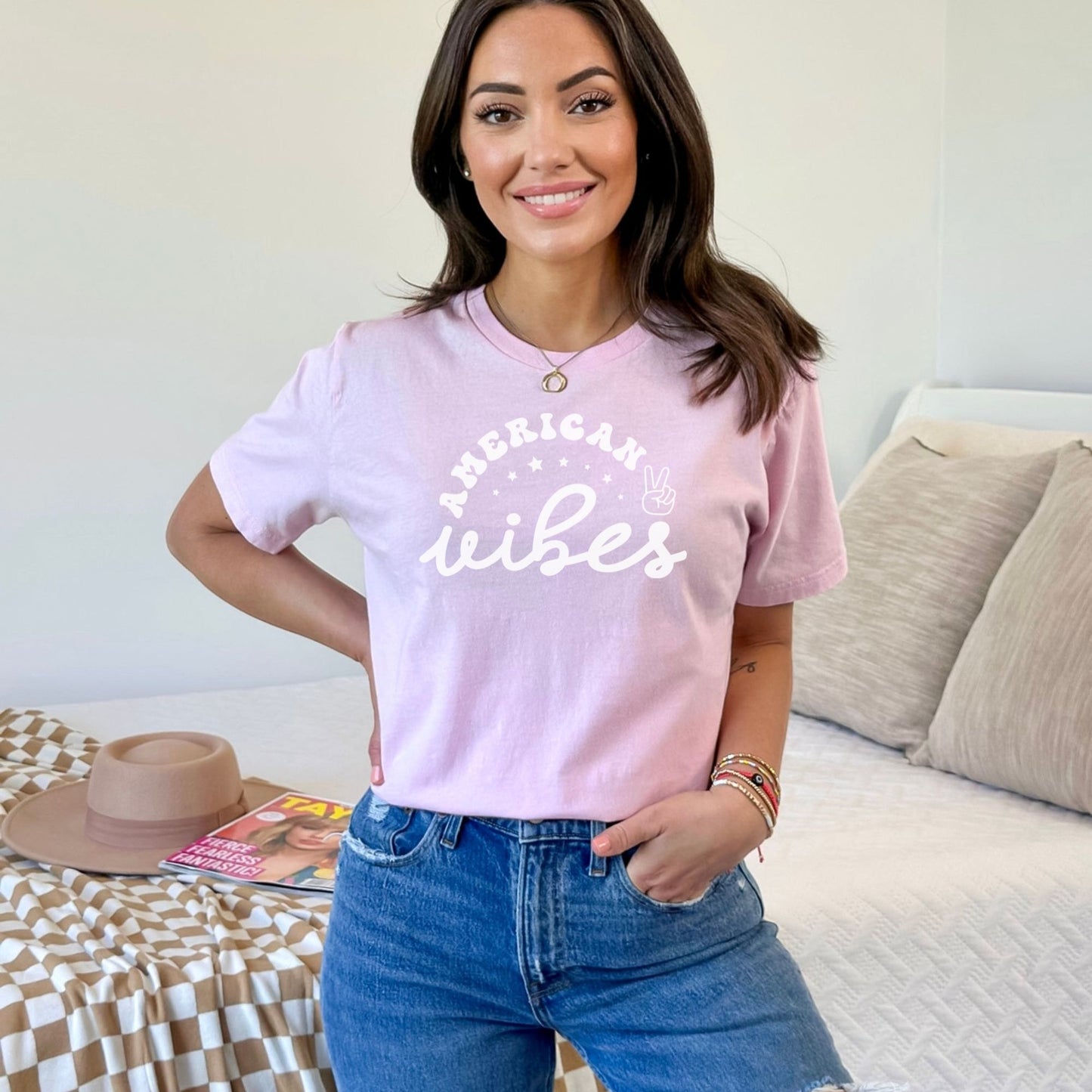 Smiling woman modeling a light pink "American Vibes" T-shirt with vintage white lettering and peace hand graphic.
