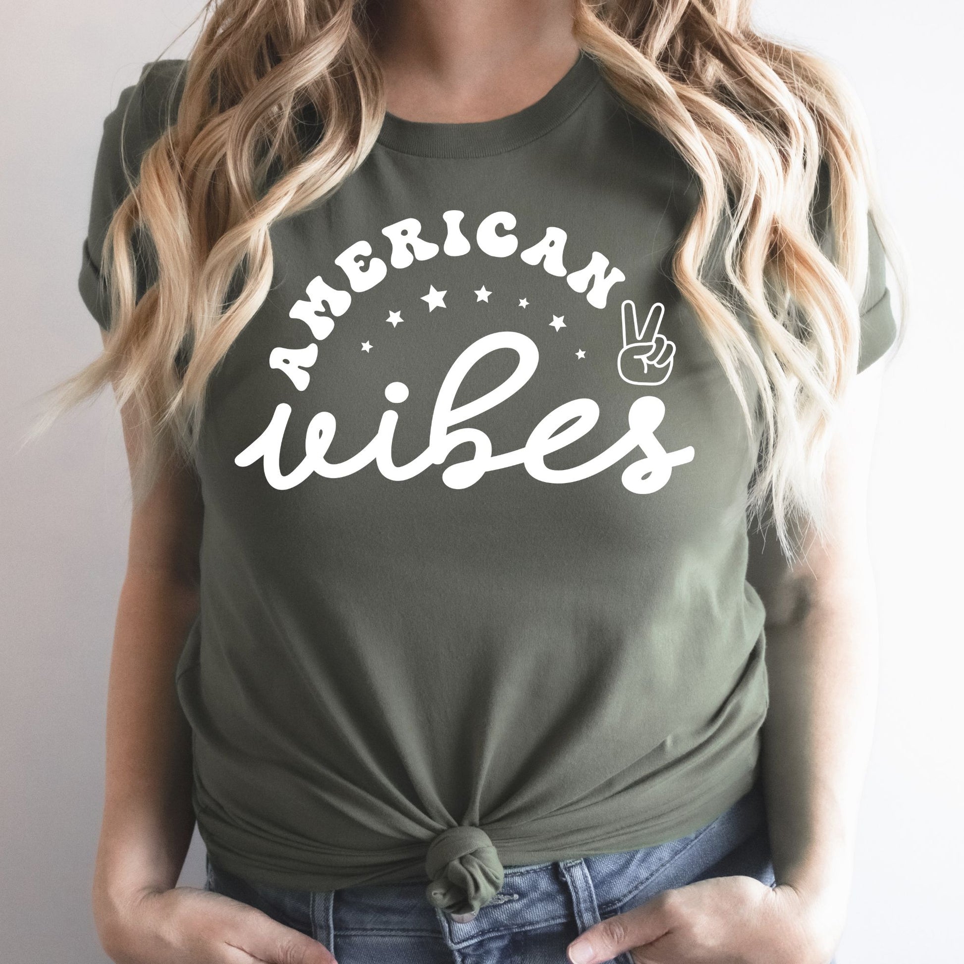 Woman wearing a military green "American Vibes" graphic tee with white retro text and stars, patriotic peace sign design.