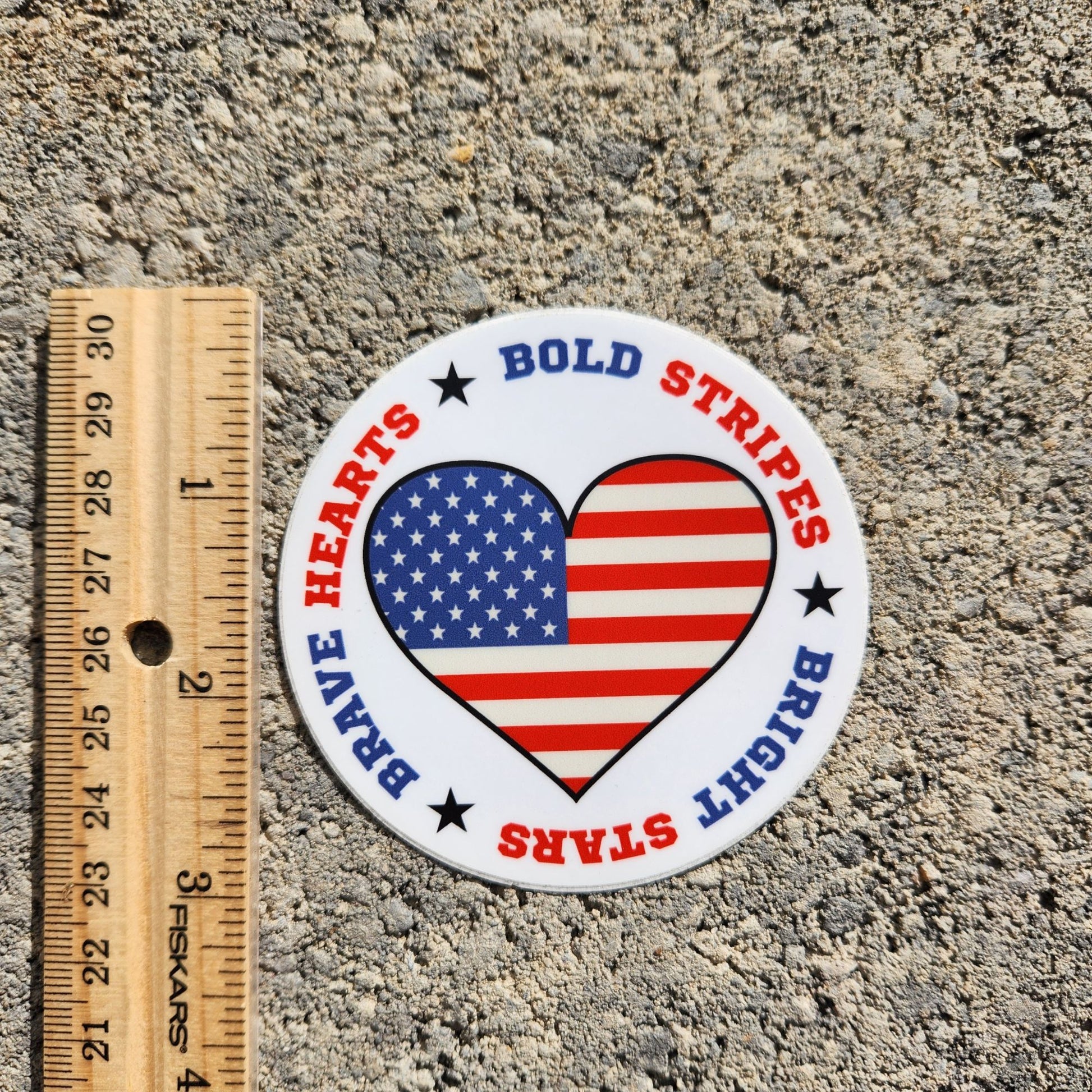 Round patriotic heart sticker with American flag design placed on concrete with ruler for scale.