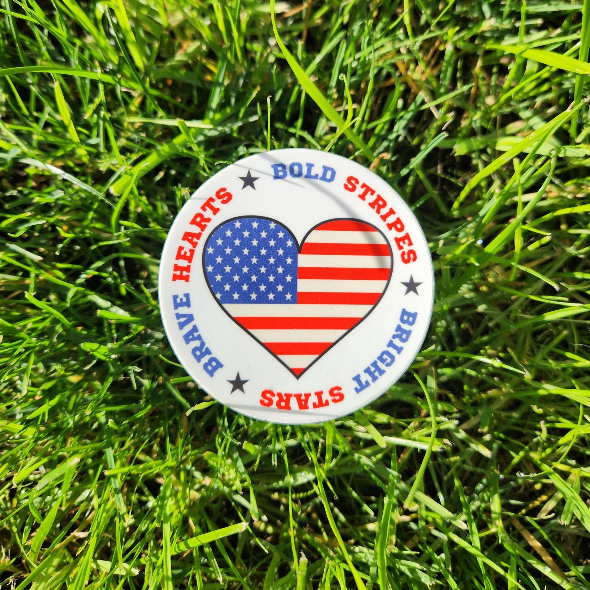 Patriotic heart flag sticker with bold stripes and stars text placed on fresh green grass.