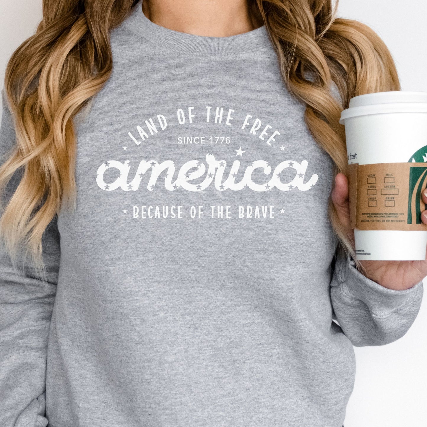 Light grey crewneck sweatshirt with patriotic "Land of the Free, America, Because of the Brave" printed on the front.