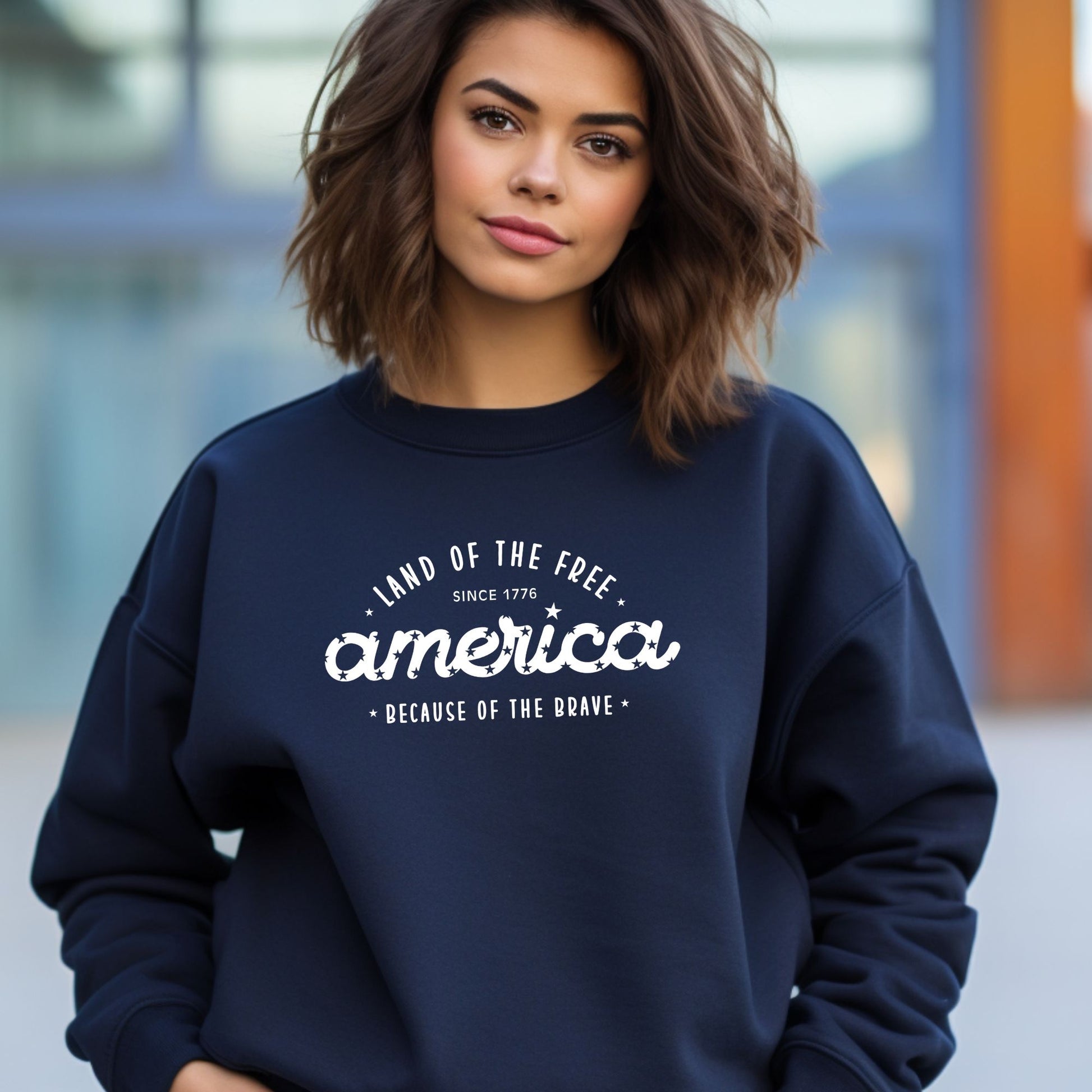 Woman wearing a navy patriotic crewneck that says "Land of the Free, America, Because of the Brave.