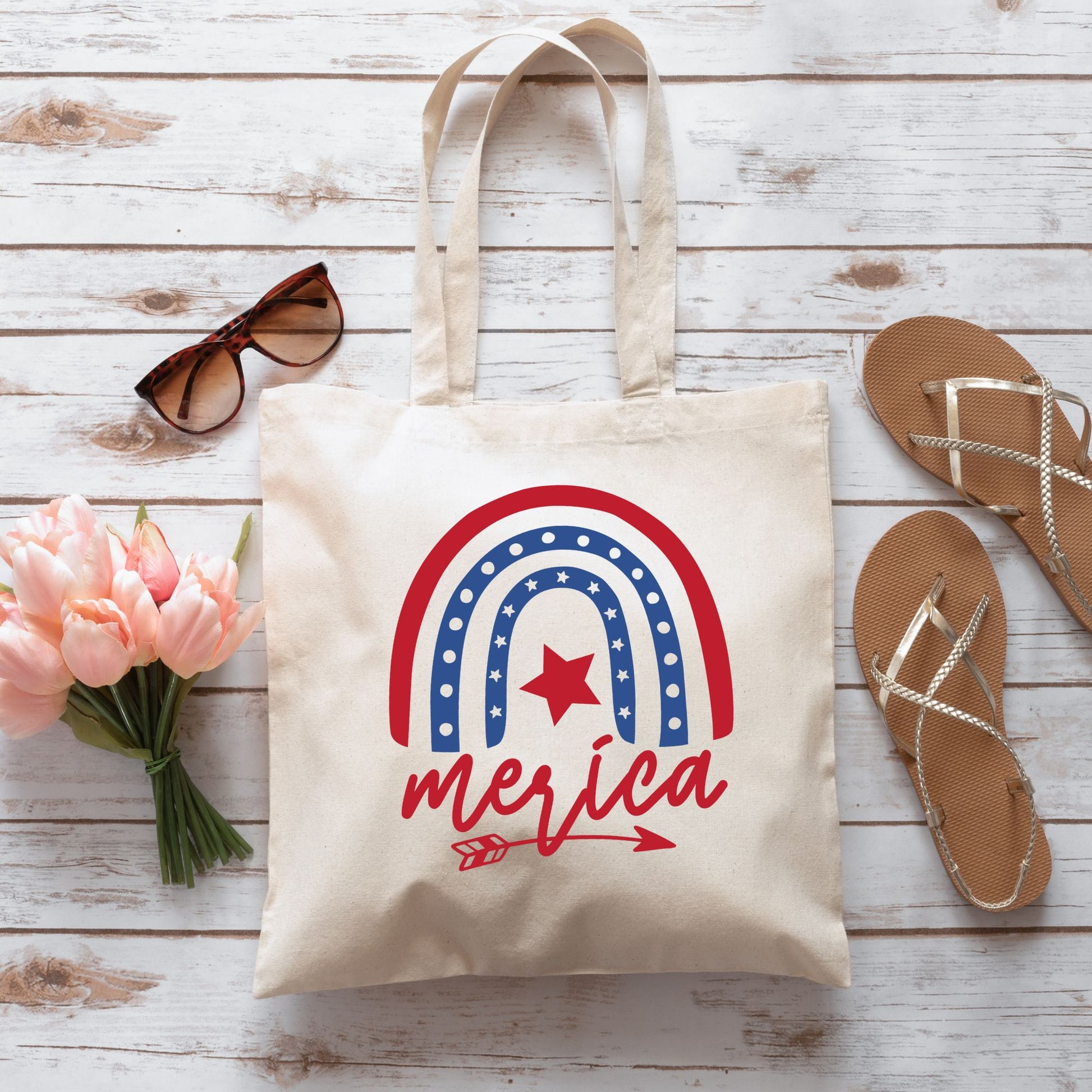 Flatlay of 'Merica tote bag with patriotic rainbow design, surrounded by sandals, sunglasses, and pink tulips on wood flooring.