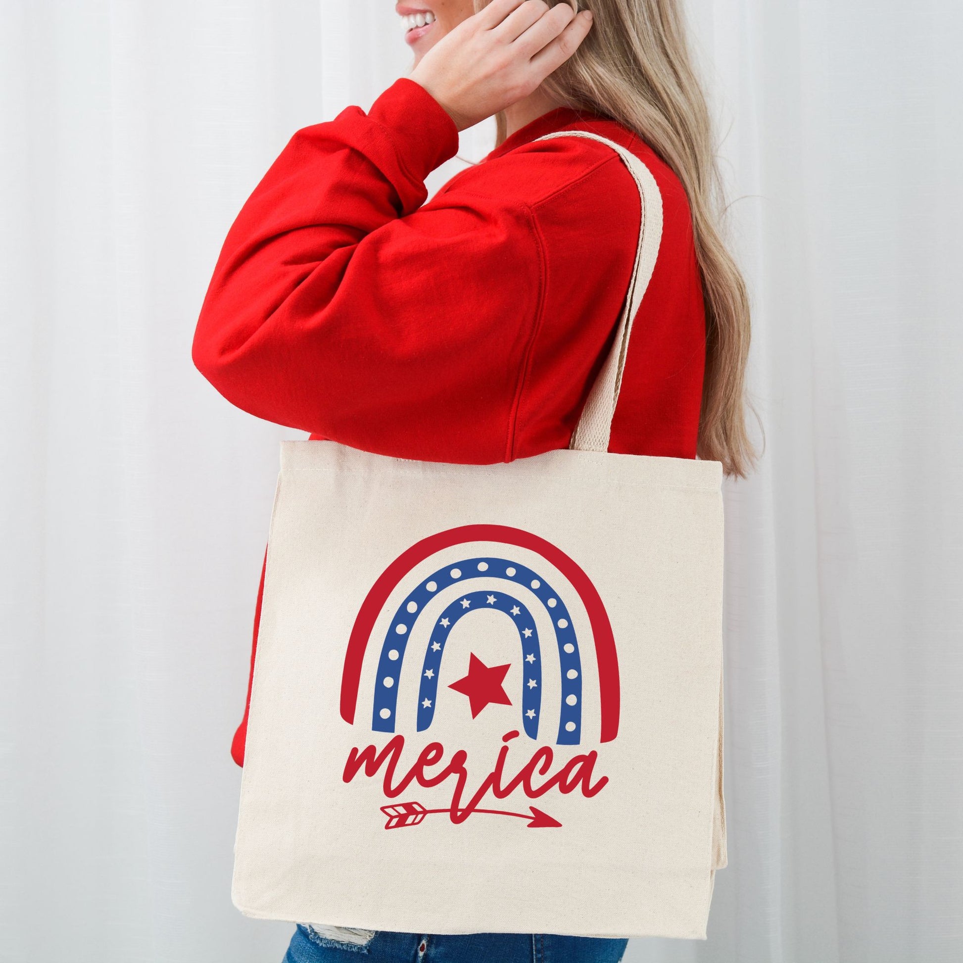 Side view of woman wearing a red sweatshirt and carrying a 'Merica patriotic tote bag with a bold rainbow and star design.