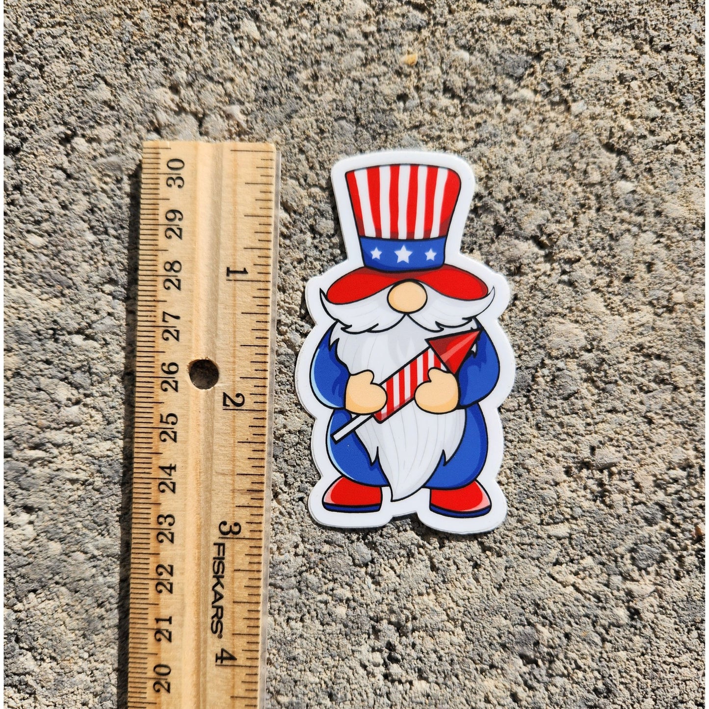 Patriotic gnome sticker next to a ruler showing its size, with a red, white, and blue design holding a firework.