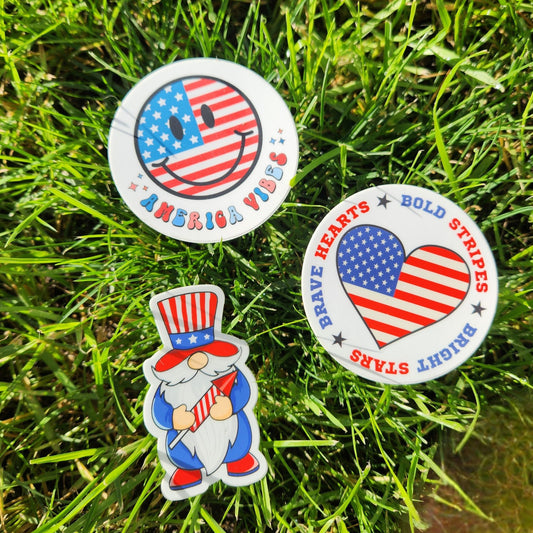 Patriotic sticker pack featuring America Vibes smiley face, red white and blue gnome, and heart-shaped Brave Hearts Bold Stripes Bright Stars design on green grass background.
