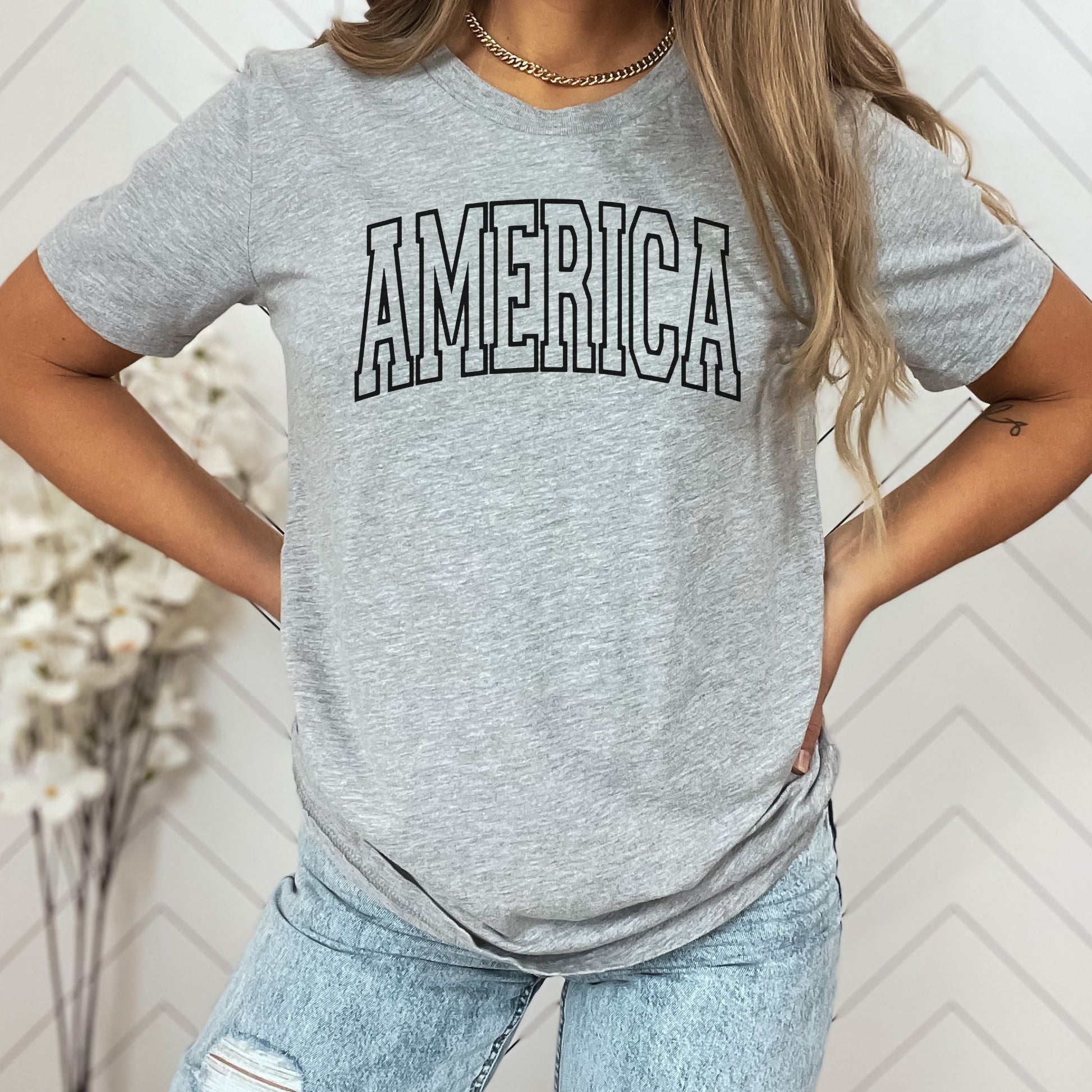 America text in arched varsity style on athletic grey women's t-shirt, modeled with light-wash jeans.