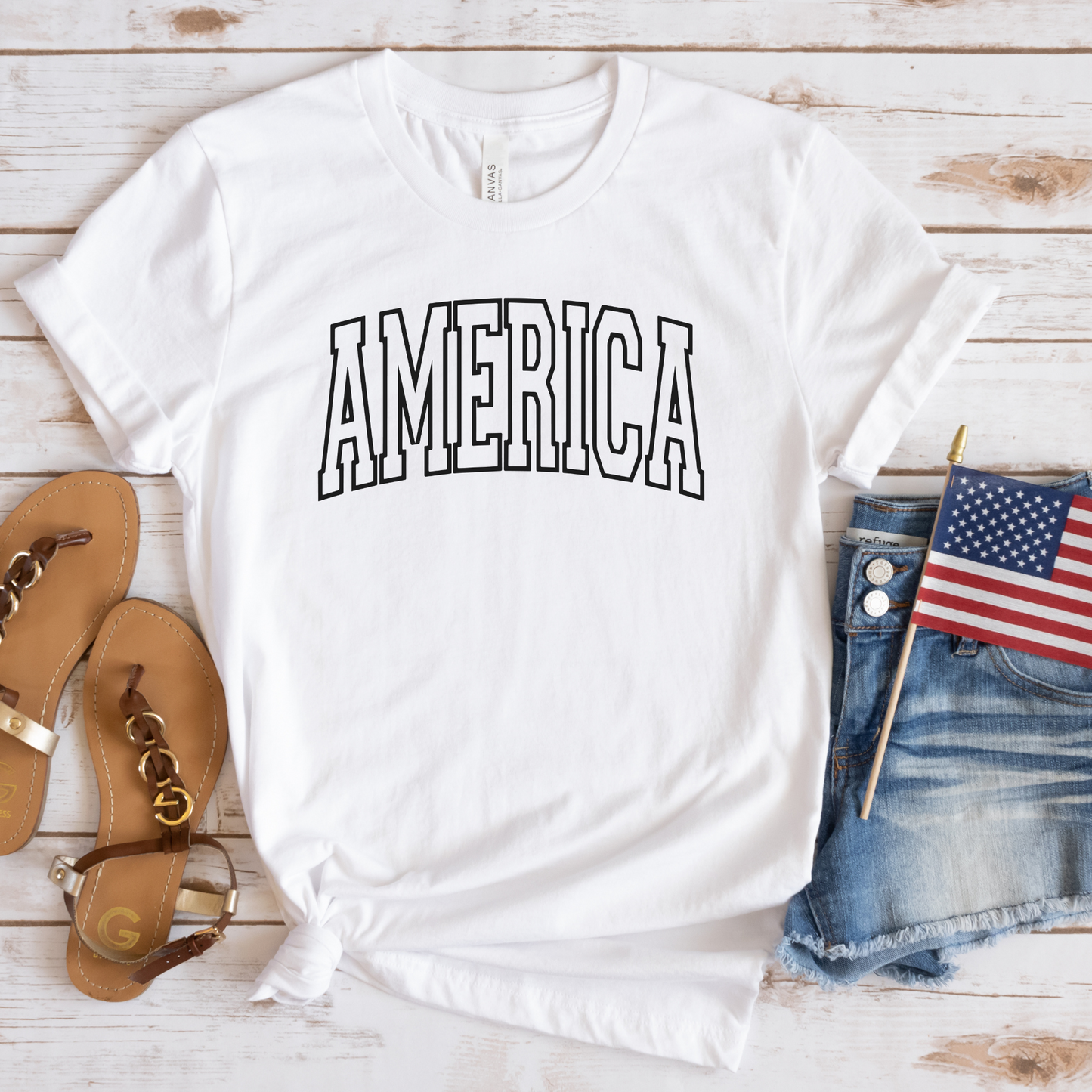 Classic America arched print on white t-shirt, flat lay styled with sandals, denim, and flag.