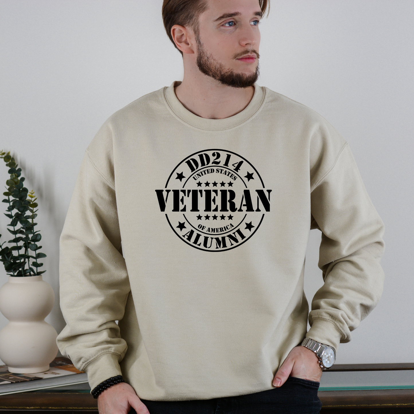 DD214 Veteran Alumni emblem on sand-colored crewneck sweatshirt, modeled indoors with minimalist decor.