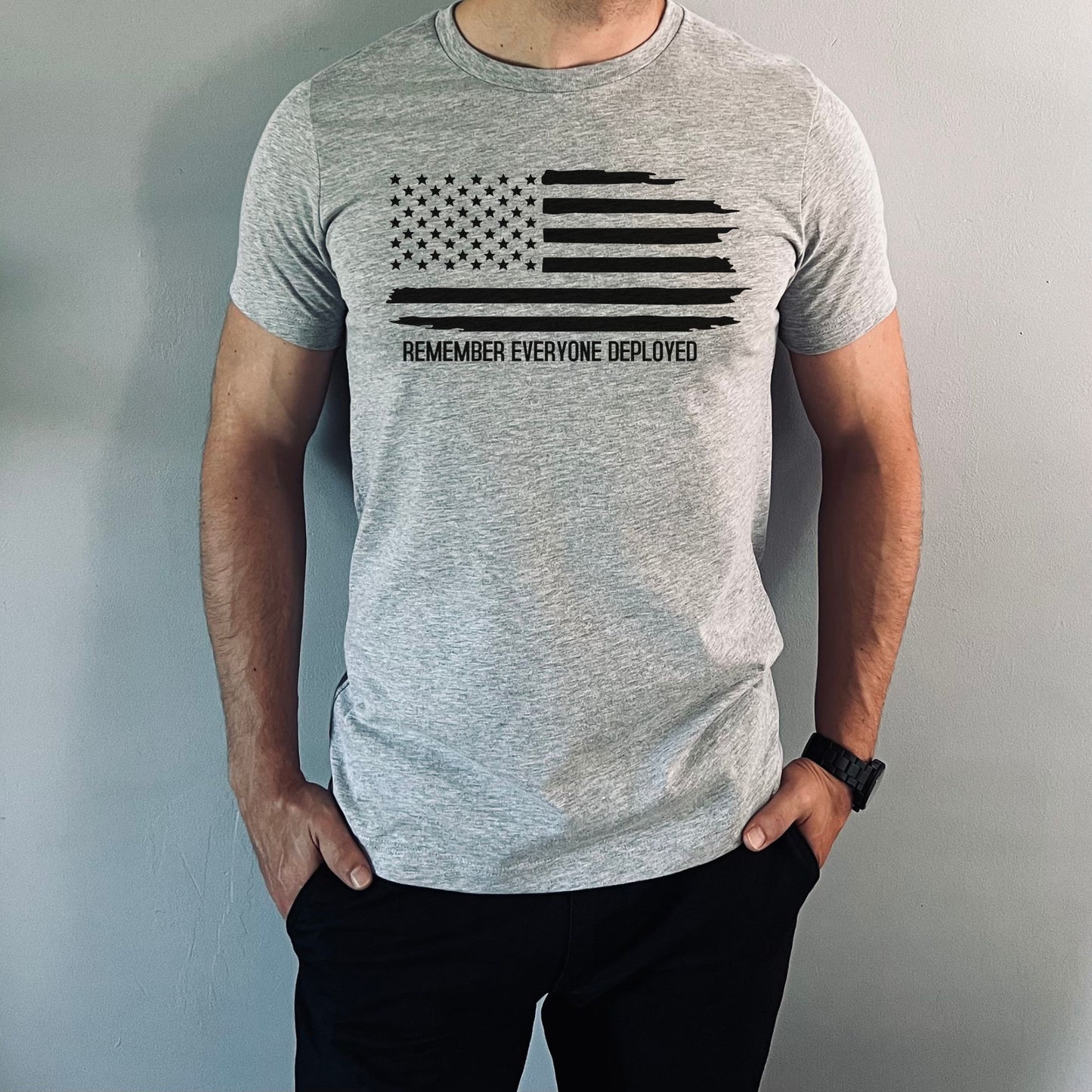 Grunge flag and Remember Everyone Deployed print on athletic grey men's t-shirt, modeled indoors.