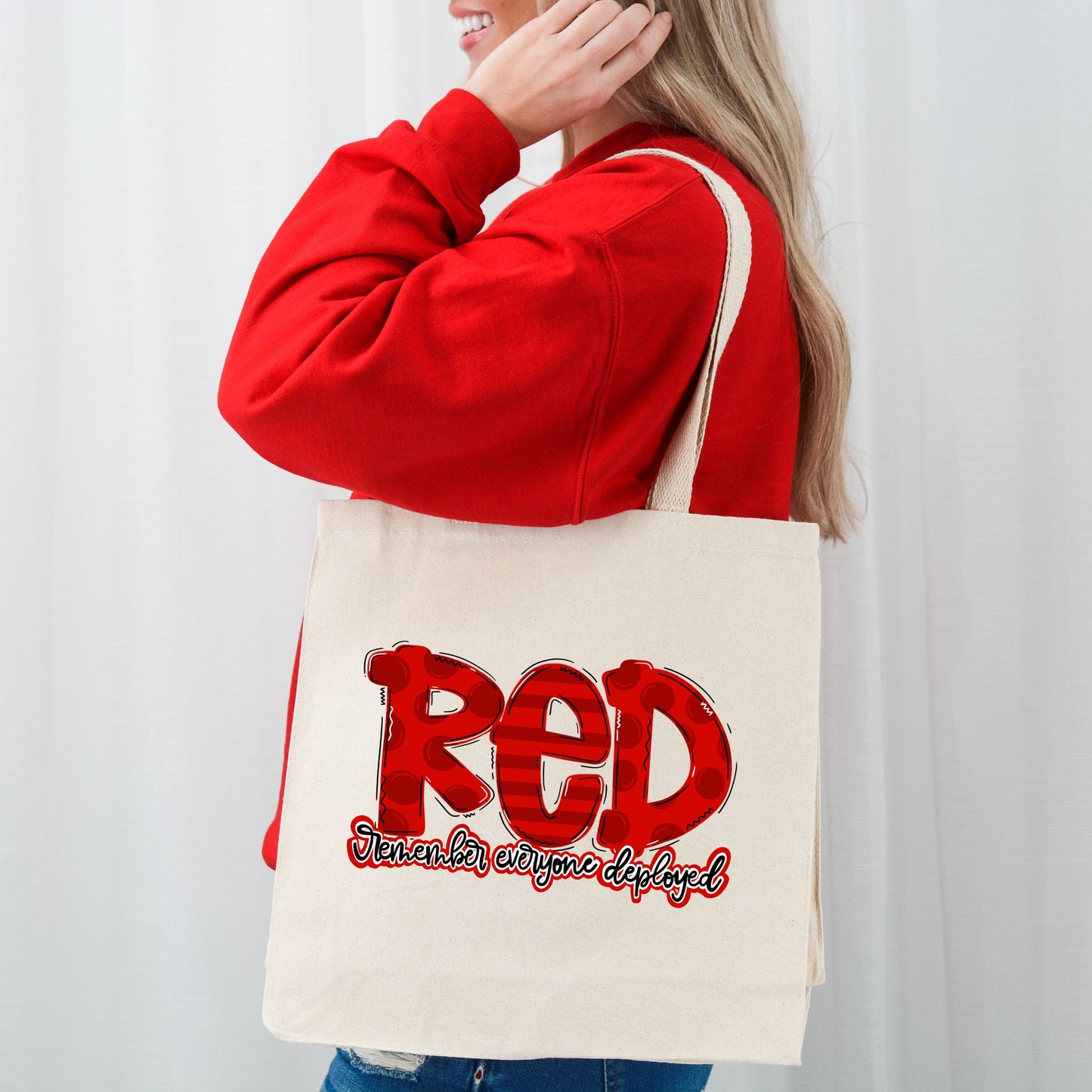 Smiling woman wearing red sweater and carrying “RED” canvas tote bag with patriotic design supporting deployed troops.