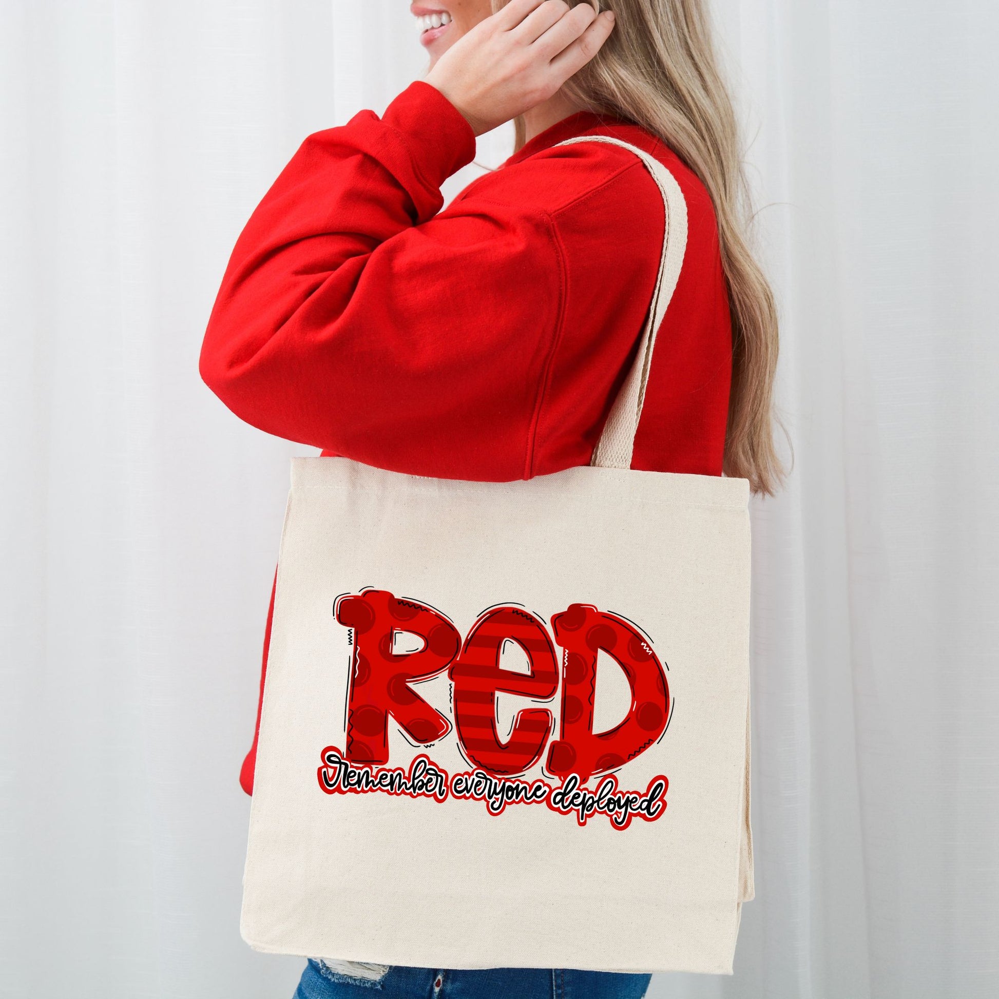 Smiling woman wearing red sweater and carrying “RED” canvas tote bag with patriotic design supporting deployed troops.